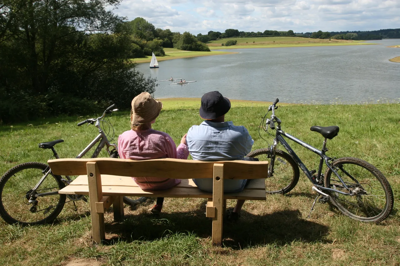 Two people on a bench overlooking Bewl Water, with a sailing boat on the water and bicycles beside them.