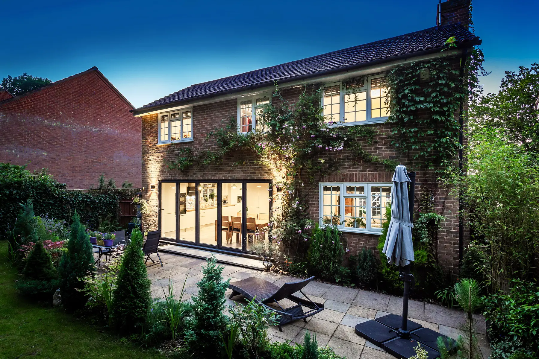 An ivy-clad brick home at twilight, bifold doors glowing warmly onto a lit terrace garden — premium twilight photography by a Kings Estates property specialist