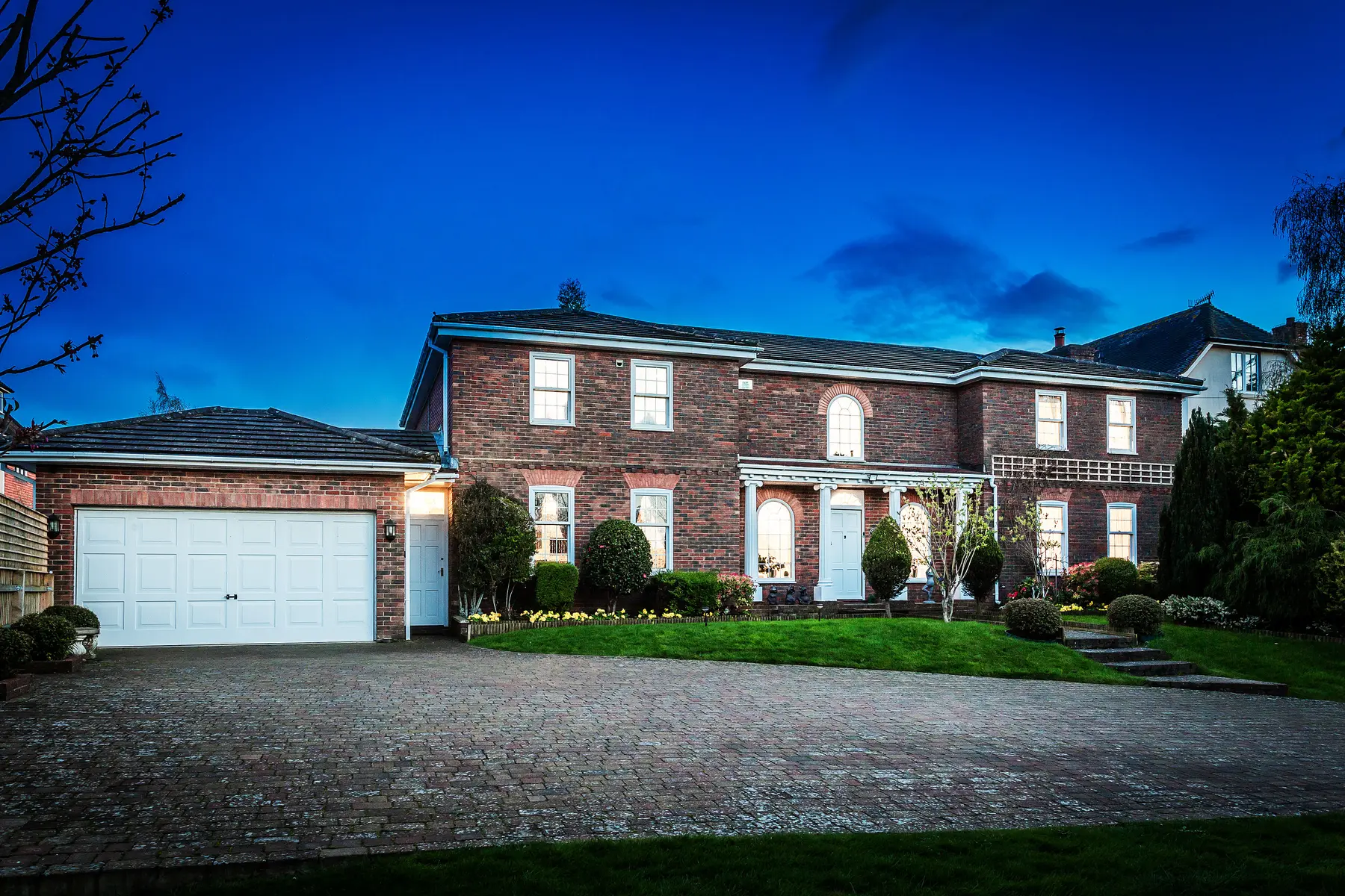 A double-fronted brick home at twilight, windows lit warmly against a deep blue sky, with spring planting along the drive — a Kings Estates home