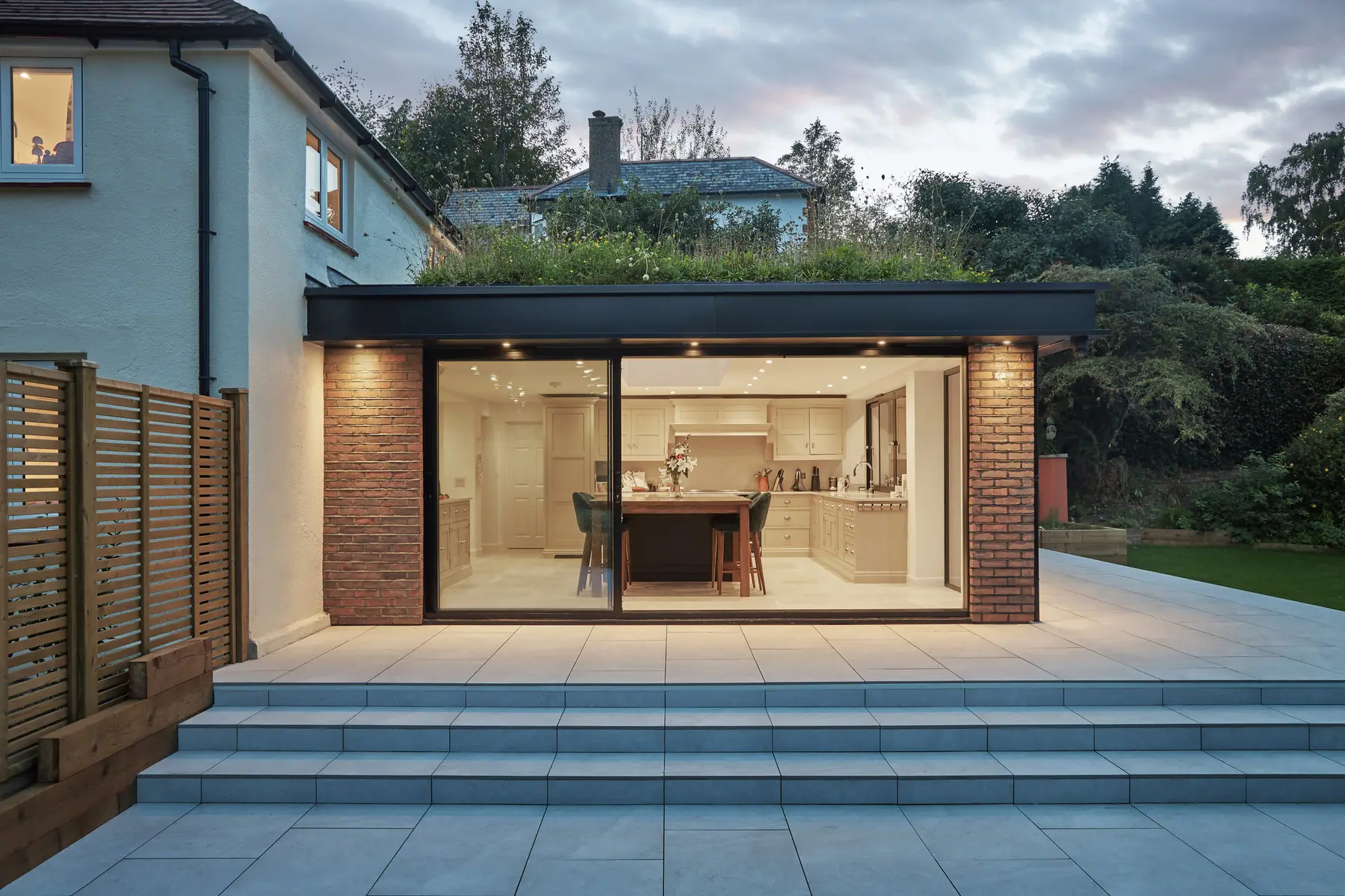 A contemporary home glowing at dusk, its open-plan kitchen visible through glass doors onto the garden — of the kind sold through Kings Estates in the Tunbridge Wells area