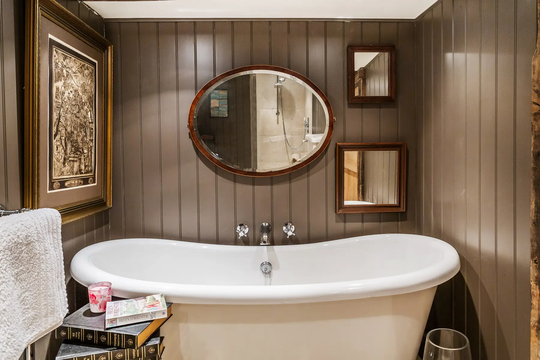 A freestanding roll-top bath beneath a panelled wall and oval mirror, styled for photography in a period home