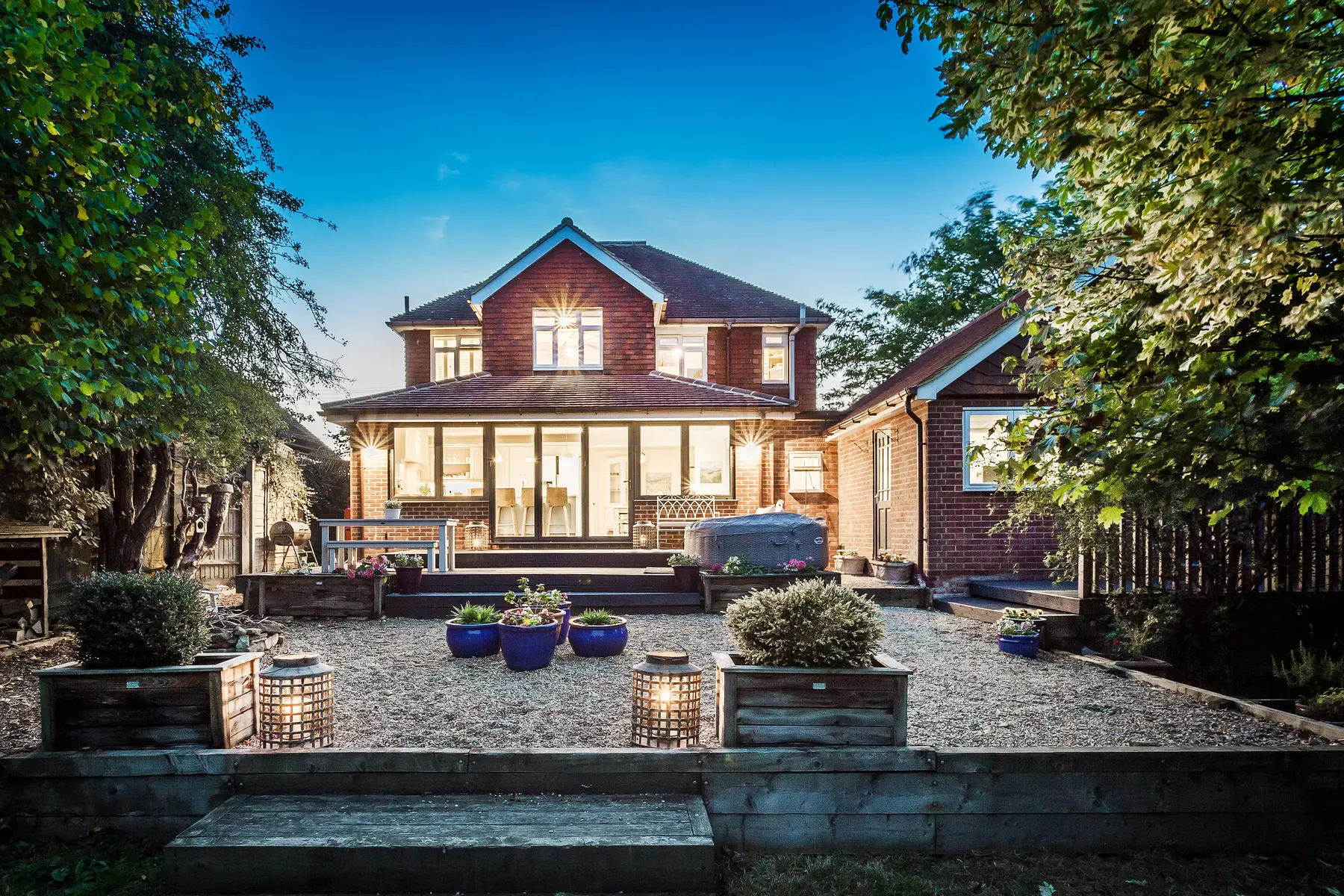 The rear of a family home glowing at dusk, every window lit above a planted terrace garden — a Kings Estates home