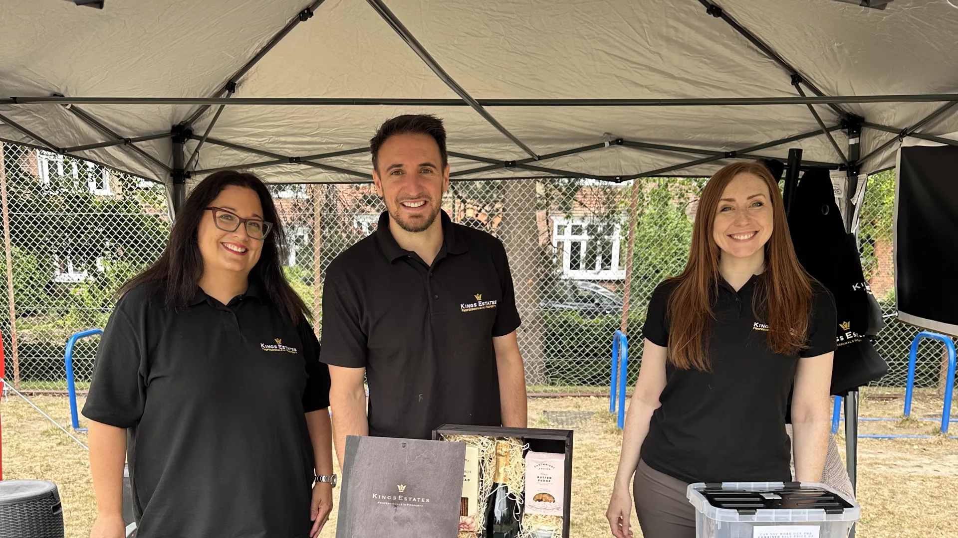 The Kings Estates team behind their stall at the Claremont Primary School Summer Fete — three colleagues in branded polo shirts under a gazebo, beside a Kings Estates table of hampers and property marketing.