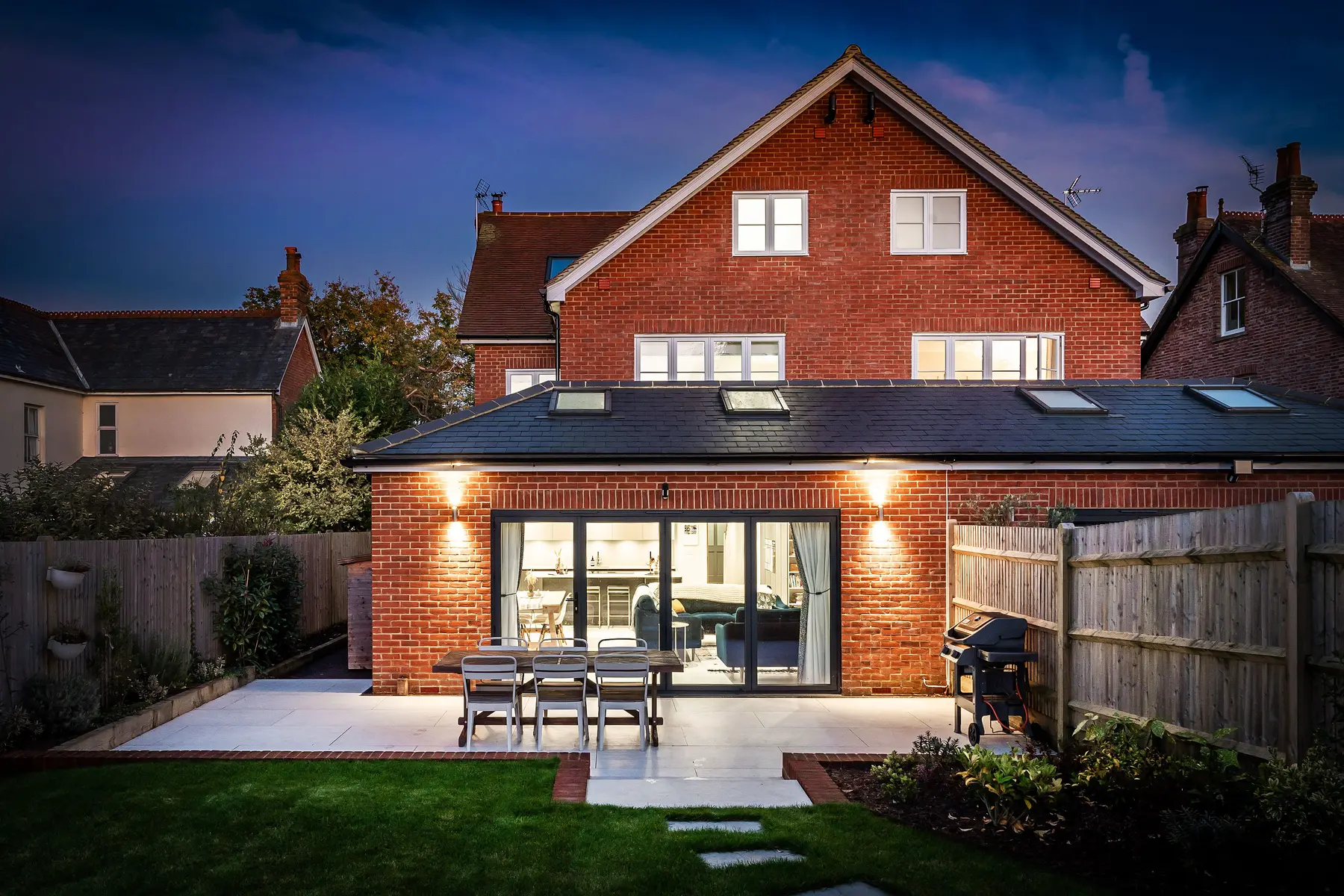A brick home at twilight with bifold doors open onto a lit terrace and dining set, every window glowing — a Kings Estates managed home